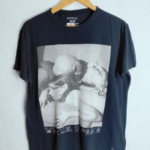 May include: Navy blue t-shirt with a black and white graphic print. The graphic features a grayscale image of people embracing, with the words "WE CUM IS PEACE" printed below. The shirt is hanging on a wooden hanger.