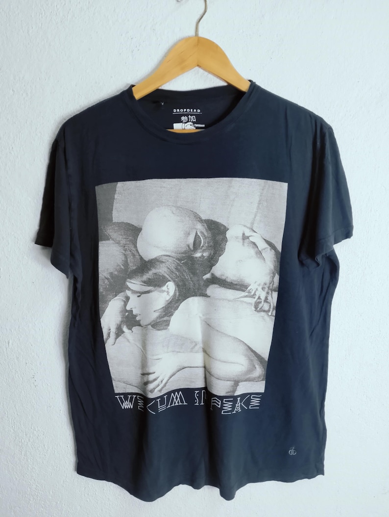 May include: Navy blue t-shirt with a black and white graphic print. The graphic features a grayscale image of people embracing, with the words "WE CUM IS PEACE" printed below. The shirt is hanging on a wooden hanger.
