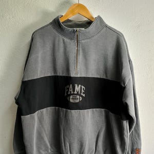 Può includere: Felpa grigia e nera con una striscia nera sul petto e la parola "FAME" in lettere bianche con un grafico di un pallone da calcio sotto. La felpa ha una cerniera sul davanti.