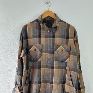 Op de afbeelding: Een geruit shirt met lange mouwen in bruin-, grijs- en oranje tinten, met een kraag met ritssluiting en twee borstzakken. Het shirt hangt aan een houten hanger tegen een witte achtergrond.