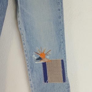 Pode incluir: Jeans de ganga azul claro com aplica&ccedil;&otilde;es decorativas. Uma aplica&ccedil;&atilde;o apresenta um desenho de sol com detalhes em laranja e amarelo, enquanto a outra &eacute; uma aplica&ccedil;&atilde;o retangular com um padr&atilde;o cinza e azul. Os jeans s&atilde;o de estilo cl&aacute;ssico.