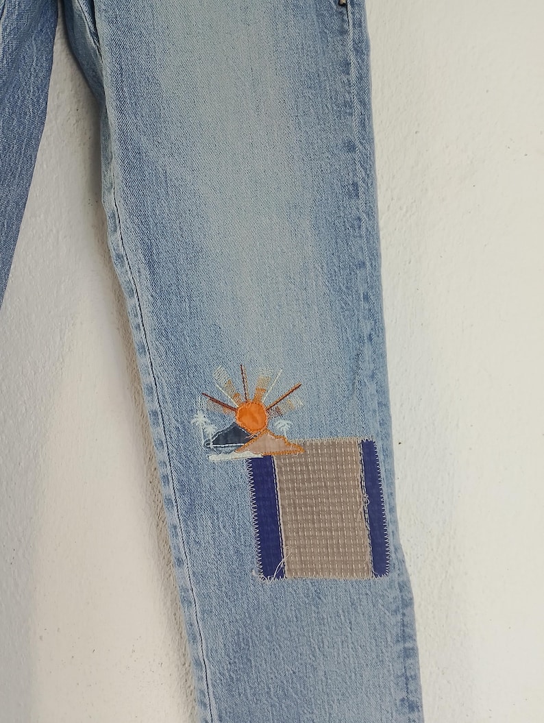 Pode incluir: Jeans de ganga azul claro com aplica&ccedil;&otilde;es decorativas. Uma aplica&ccedil;&atilde;o apresenta um desenho de sol com detalhes em laranja e amarelo, enquanto a outra &eacute; uma aplica&ccedil;&atilde;o retangular com um padr&atilde;o cinza e azul. Os jeans s&atilde;o de estilo cl&aacute;ssico.