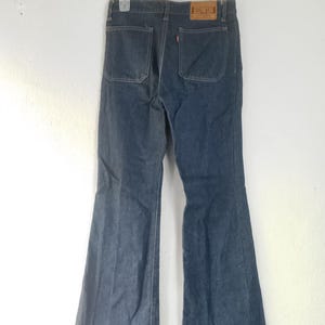 Puede incluir: Vaqueros de mezclilla azul oscuro con dise&ntilde;o de pernera acampanada. Los vaqueros tienen dos bolsillos traseros y un parche de cuero marr&oacute;n con la marca "EDWIN". Los vaqueros est&aacute;n colgados de una percha blanca.