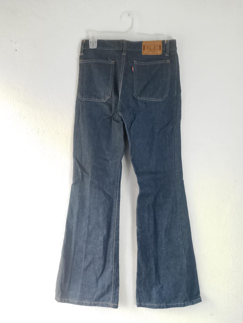 Puede incluir: Vaqueros de mezclilla azul oscuro con dise&ntilde;o de pernera acampanada. Los vaqueros tienen dos bolsillos traseros y un parche de cuero marr&oacute;n con la marca "EDWIN". Los vaqueros est&aacute;n colgados de una percha blanca.