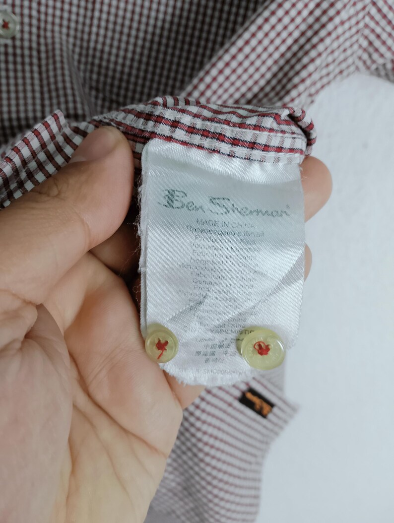 K&ouml;nnte beinhalten: Ein Ben Sherman Hemd mit rot-wei&szlig;em Vichy-Karomuster. Das innere Pflegeetikett ist sichtbar und zeigt den Markennamen, das Herkunftsland sowie zwei Ersatzkn&ouml;pfe, die am unteren Ende des wei&szlig;en Stoffetiketts befestigt sind.