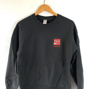 Puede incluir: Una sudadera negra de cuello redondo con un peque&ntilde;o logotipo de Coca-Cola en rojo y blanco en el pecho. El logotipo va acompa&ntilde;ado de texto rojo. La sudadera est&aacute; colgada de una percha de madera.