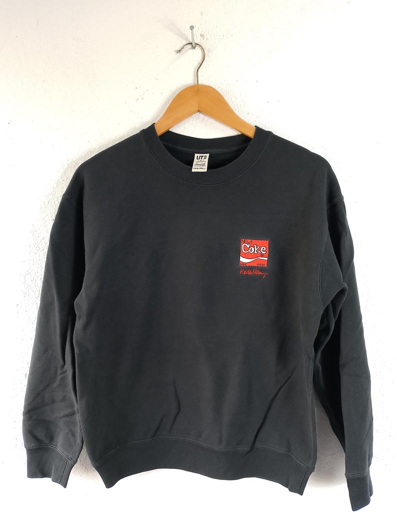 Puede incluir: Una sudadera negra de cuello redondo con un peque&ntilde;o logotipo de Coca-Cola en rojo y blanco en el pecho. El logotipo va acompa&ntilde;ado de texto rojo. La sudadera est&aacute; colgada de una percha de madera.