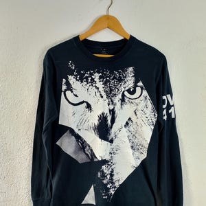 Pode incluir: Camiseta preta de manga comprida com um grande gr&aacute;fico branco do rosto de uma coruja. O design &eacute; geom&eacute;trico e abstrato. A manga tem o texto "OV 41" impresso. A camiseta est&aacute; pendurada em um cabide de madeira.