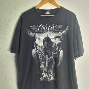May include: Dark grey t-shirt with a detailed graphic of a Native American figure, a buffalo skull, and the word "Chiefbon" in a decorative font. The design is in shades of white and grey.