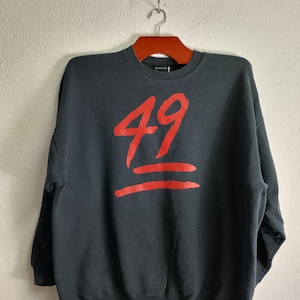May include: A dark grey sweatshirt featuring the number "49" in red, with two red horizontal lines beneath. The garment is suspended on a wooden hanger against a plain white wall.