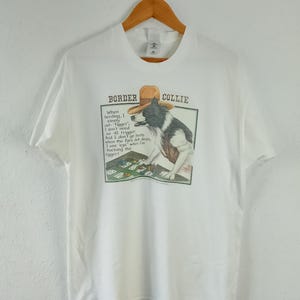 Peut inclure: T-shirt blanc avec un motif de Border Collie portant un chapeau et jouant aux cartes. Le texte dit : "When herding, I simply out-Figger..." Le t-shirt est suspendu à un cintre en bois.