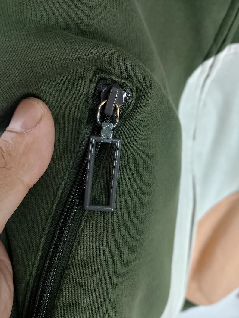 May include: Close-up of a dark green jacket with a black zipper. The zipper pull is a rectangular shape. The jacket has a contrasting white and peach-colored panel.