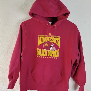 May include: Maroon hooded sweatshirt featuring the University of Minnesota Golden Gophers logo. The design includes the word "Minnesota" in gold lettering, a football helmet graphic, and the words "Golden Gophers". The sweatshirt has a front pocket and a drawstring hood.