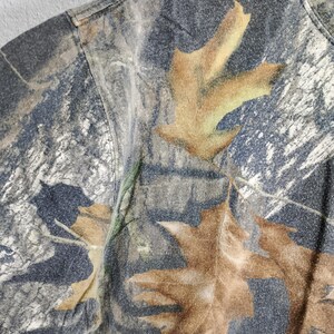 May include: Close-up of a camouflage t-shirt featuring a pattern of brown leaves and branches on a gray and green background. The short-sleeved shirt has a crew neck and is made of a soft, textured fabric.