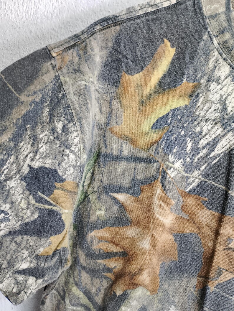 May include: Close-up of a camouflage t-shirt featuring a pattern of brown leaves and branches on a gray and green background. The short-sleeved shirt has a crew neck and is made of a soft, textured fabric.