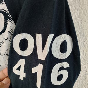Pode incluir: Manga de camisa preta com texto branco. O texto "OVO 416" est&aacute; escrito em negrito, com um efeito desgastado. O tecido parece macio e o desenho &eacute; impresso na manga.