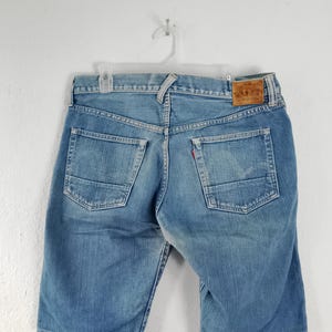 Puede incluir: Pantalones cortos vaqueros azul claro con un diseño clásico de cinco bolsillos. Los pantalones cortos tienen un parche de cuero marrón en la cintura trasera y una etiqueta roja en el bolsillo trasero. Colgados en una percha blanca.