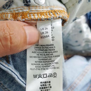 Pode incluir: Close-up de uma etiqueta de roupa de ganga azul clara. A etiqueta exibe informa&ccedil;&otilde;es de tamanho: US 26, UK 26 e 165/68A. As instru&ccedil;&otilde;es de lavagem tamb&eacute;m s&atilde;o impressas na etiqueta.