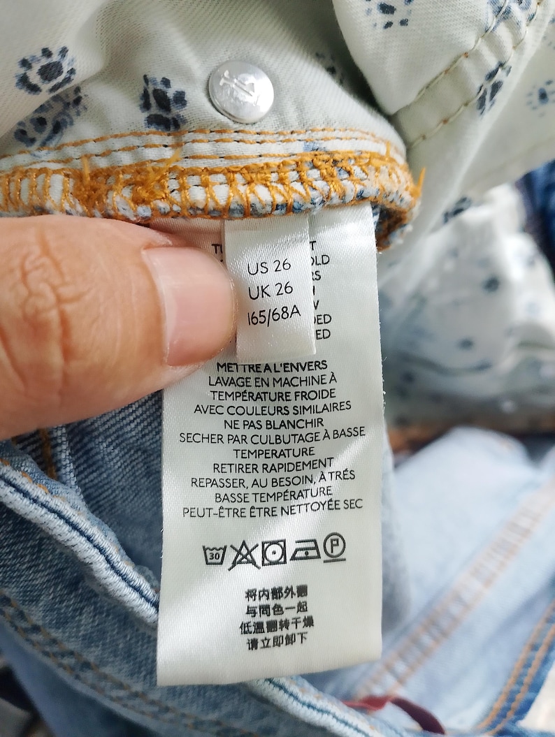 Pode incluir: Close-up de uma etiqueta de roupa de ganga azul clara. A etiqueta exibe informa&ccedil;&otilde;es de tamanho: US 26, UK 26 e 165/68A. As instru&ccedil;&otilde;es de lavagem tamb&eacute;m s&atilde;o impressas na etiqueta.