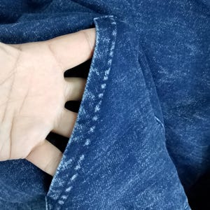 Pode incluir: Close-up de tecido denim azul escuro, mostrando a textura e as costuras de um bolso. O tecido est&aacute; dobrado, revelando o interior do bolso. As costuras s&atilde;o brancas, contrastando com o denim azul escuro.