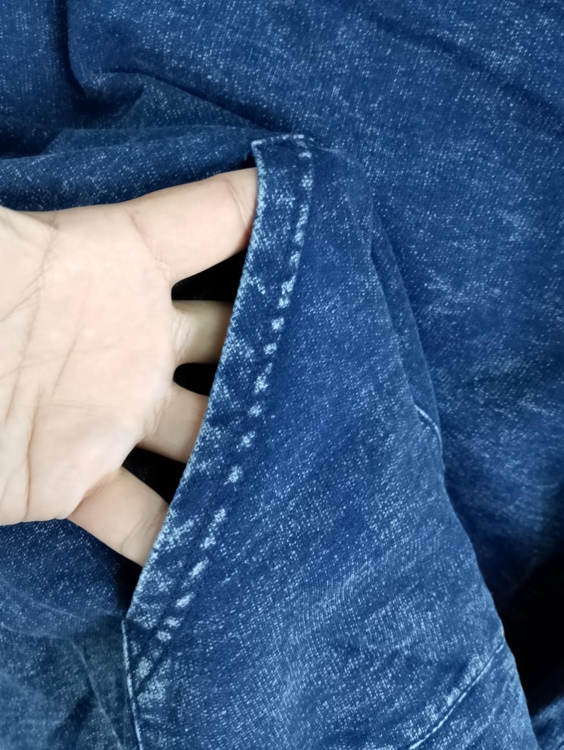 Pode incluir: Close-up de tecido denim azul escuro, mostrando a textura e as costuras de um bolso. O tecido est&aacute; dobrado, revelando o interior do bolso. As costuras s&atilde;o brancas, contrastando com o denim azul escuro.