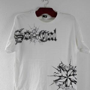 Könnte beinhalten: Weißes T-Shirt mit schwarzem Grafiktext und Sternenmuster. Der Text lautet "So Cal" in einer gotischen Schriftart über einem Sternenmuster. Ein sternförmiges Design mit dem Text "So Cal Republic" ist ebenfalls sichtbar.