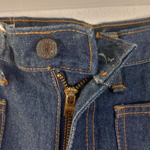 Puede incluir: Primer plano de unos vaqueros de mezclilla azules con cremallera y cierre de bot&oacute;n de lat&oacute;n. Los vaqueros tienen costuras naranjas y detalles visibles de la marca Edwin. La cremallera est&aacute; parcialmente abierta, mostrando la textura y construcci&oacute;n de la tela.