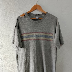 May include: A grey t-shirt with a faded blue, red and white striped band across the chest. The band has a faded blue logo with the word "Stussy" above it.