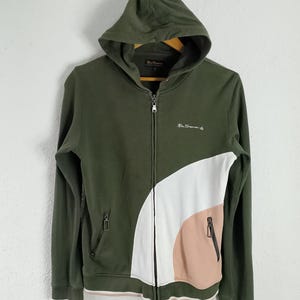 May include: Olive green zip-up hoodie with a color block design in white and peach. Features a hood, two front pockets, and the brand name embroidered on the chest. The cuffs and hem have a thin pink and white stripe.