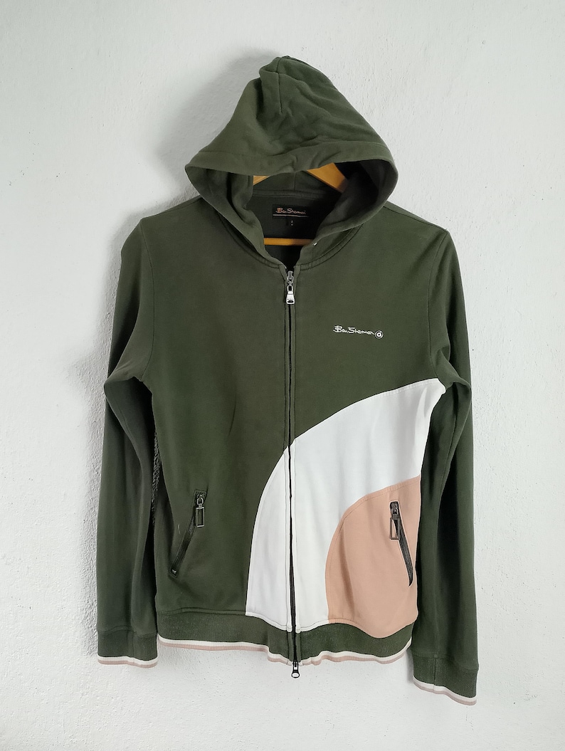 May include: Olive green zip-up hoodie with a color block design in white and peach. Features a hood, two front pockets, and the brand name embroidered on the chest. The cuffs and hem have a thin pink and white stripe.
