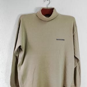 May include: A beige long-sleeve turtleneck shirt with a vertical ribbed texture. The shirt features the word DOCKERS embroidered in dark blue on the upper left chest and a small rectangular brand tag near the bottom hem on the front.