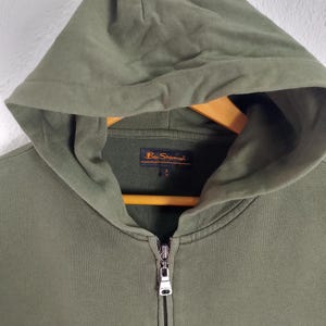 May include: Olive green zip-up hoodie with a hood and a small logo on the chest. The hoodie has a full zipper closure and a drawstring hood. The brand name "Ben Sherman" is visible on the chest and the label.