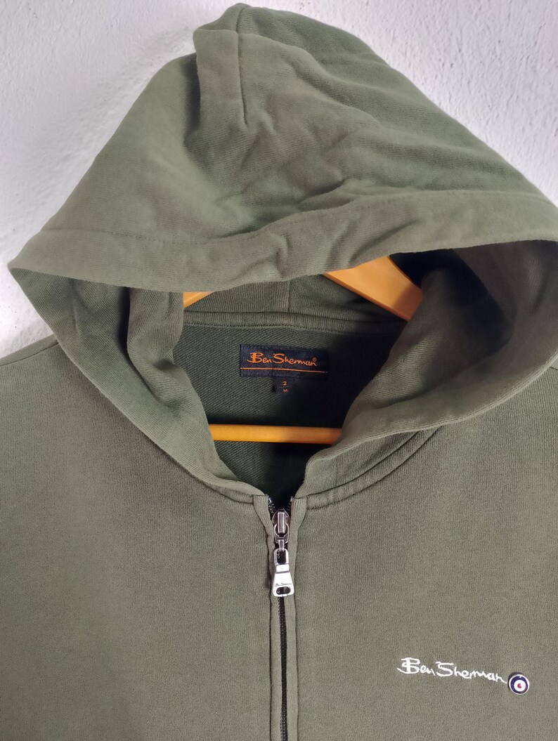 May include: Olive green zip-up hoodie with a hood and a small logo on the chest. The hoodie has a full zipper closure and a drawstring hood. The brand name "Ben Sherman" is visible on the chest and the label.