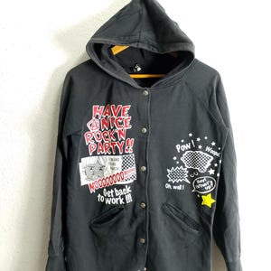 May include: Black hooded sweatshirt with a button-up front. The sweatshirt features graphic text and illustrations in red, white, and yellow. Text includes "Have a Nice Rock'n Party!!" and comic book style speech bubbles.