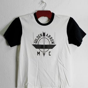 May include: White t-shirt with black sleeves and a black ringer neck. The shirt has a black and white logo that says "Golden Arrow MC" with a stylized arrow and wings.