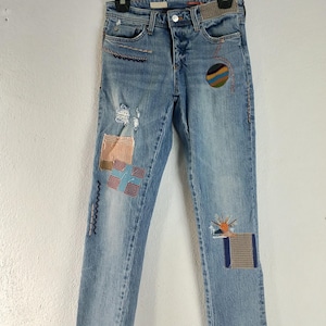 Pode incluir: Jeans de ganga azul claro com aplica&ccedil;&otilde;es decorativas e detalhes desgastados. As aplica&ccedil;&otilde;es incluem padr&otilde;es geom&eacute;tricos, um desenho de sol e um desenho circular. As cal&ccedil;as de ganga est&atilde;o penduradas num cabide preto.