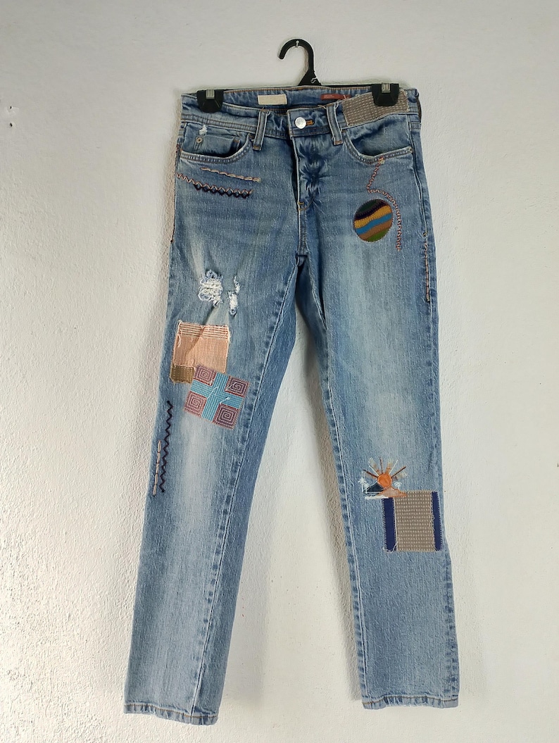 Pode incluir: Jeans de ganga azul claro com aplica&ccedil;&otilde;es decorativas e detalhes desgastados. As aplica&ccedil;&otilde;es incluem padr&otilde;es geom&eacute;tricos, um desenho de sol e um desenho circular. As cal&ccedil;as de ganga est&atilde;o penduradas num cabide preto.