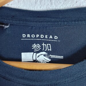 May include: Navy blue t-shirt with the text "DROPDEAD" above Japanese characters and a graphic of a hand holding a weapon. The shirt is hanging on a wooden hanger.