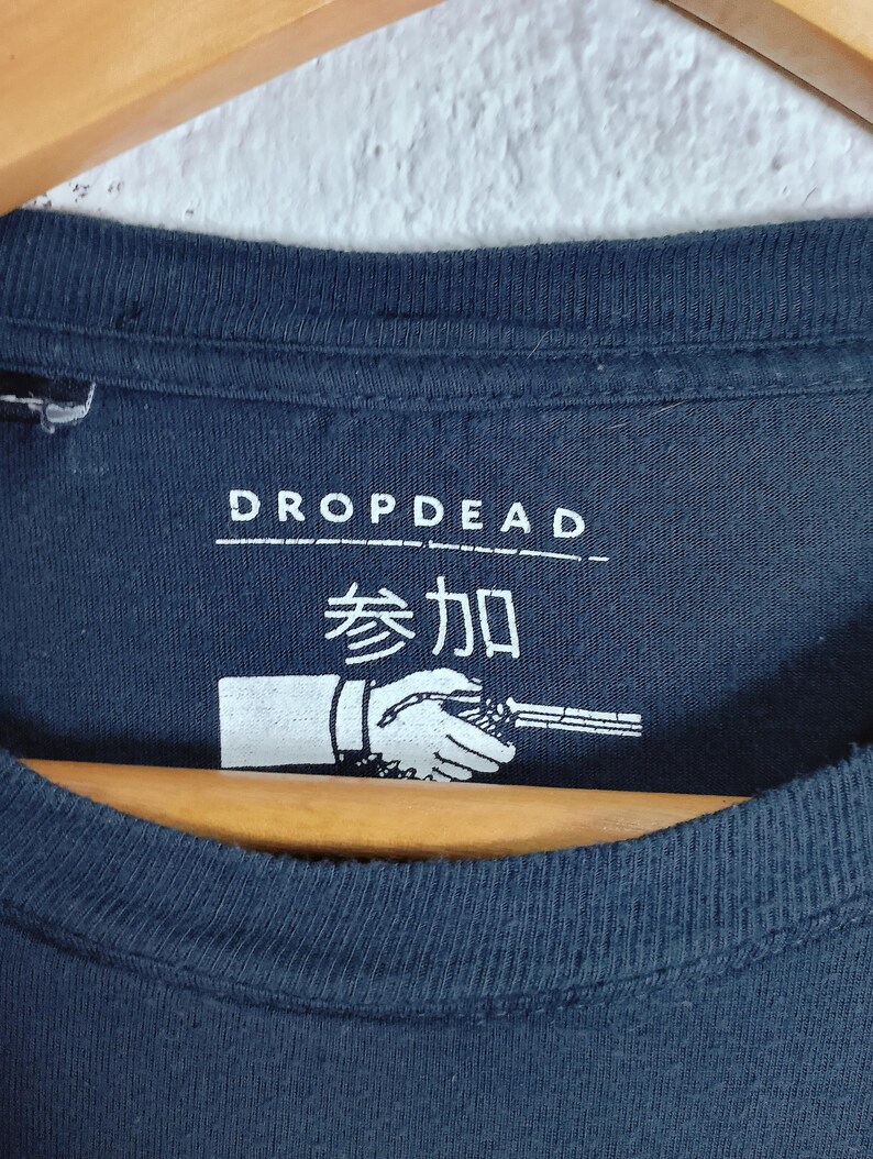 May include: Navy blue t-shirt with the text "DROPDEAD" above Japanese characters and a graphic of a hand holding a weapon. The shirt is hanging on a wooden hanger.