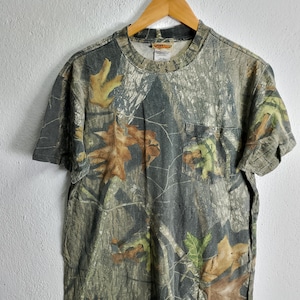 May include: A short-sleeved camouflage t-shirt with a pocket on the chest. The shirt features a pattern of brown, green, and tan leaves and branches. The shirt is hanging on a wooden hanger.