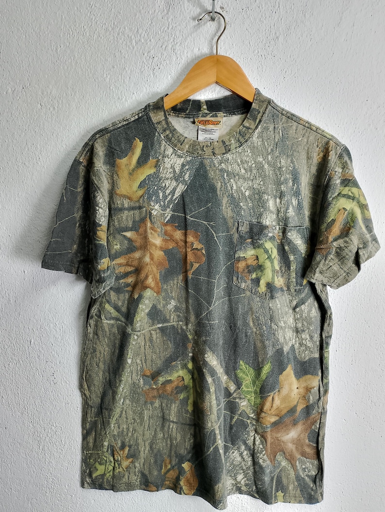 May include: A short-sleeved camouflage t-shirt with a pocket on the chest. The shirt features a pattern of brown, green, and tan leaves and branches. The shirt is hanging on a wooden hanger.
