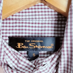 K&ouml;nnte beinhalten: Ein Hemd mit Knopfleiste und einem kleinkarierten Muster in Kastanienbraun und Wei&szlig;. Am inneren Kragen ist ein schwarzes Stoffetikett mit dem goldenen Schriftzug THE ORIGINAL Ben Sherman und einem kleinen Etikett darunter f&uuml;r die Gr&ouml;&szlig;e S angebracht.
