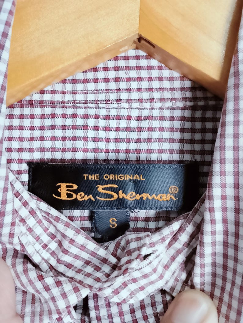 K&ouml;nnte beinhalten: Ein Hemd mit Knopfleiste und einem kleinkarierten Muster in Kastanienbraun und Wei&szlig;. Am inneren Kragen ist ein schwarzes Stoffetikett mit dem goldenen Schriftzug THE ORIGINAL Ben Sherman und einem kleinen Etikett darunter f&uuml;r die Gr&ouml;&szlig;e S angebracht.