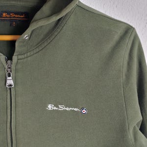 May include: Olive green zip-up hoodie with a white panel at the bottom. Features the Ben Sherman logo embroidered in white on the chest. The hoodie has a drawstring hood and a full-length zipper.