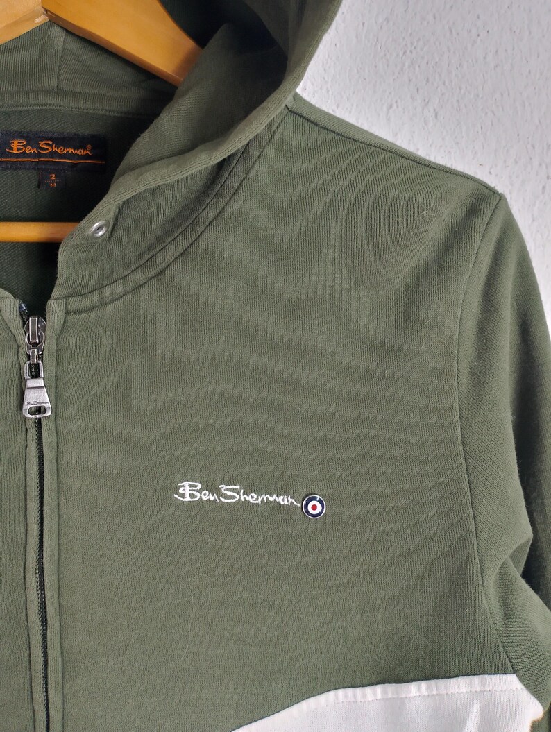 May include: Olive green zip-up hoodie with a white panel at the bottom. Features the Ben Sherman logo embroidered in white on the chest. The hoodie has a drawstring hood and a full-length zipper.