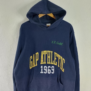Pode incluir: Camisola com capuz azul marinho com o texto "GAP ATHLETIC 1969" em amarelo e branco. A camisola também tem o texto "S.F. Calif." em verde. Possui um bolso frontal e um capuz com cordão.