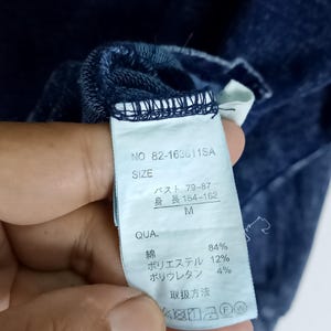 Pode incluir: Close-up de uma etiqueta de roupa azul escuro com texto japon&ecirc;s. A etiqueta inclui o tamanho, a composi&ccedil;&atilde;o do material e as instru&ccedil;&otilde;es de cuidado. A roupa &eacute; feita de algod&atilde;o, poli&eacute;ster e poliuretano.