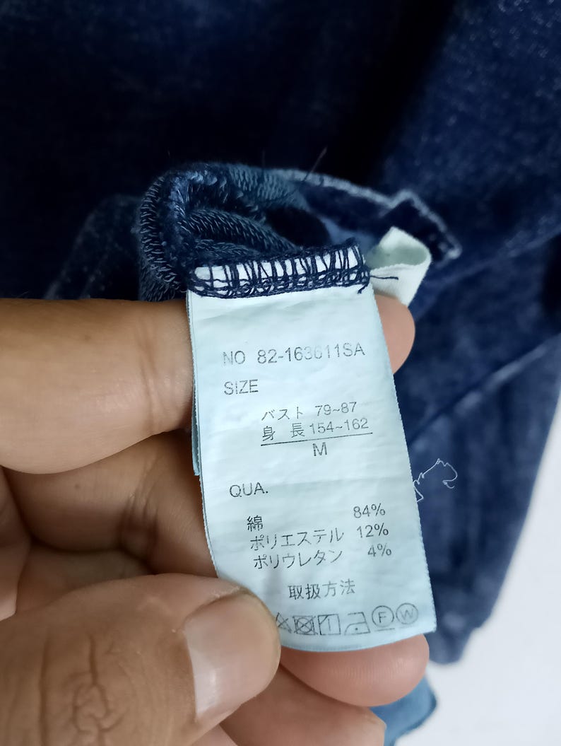 Pode incluir: Close-up de uma etiqueta de roupa azul escuro com texto japon&ecirc;s. A etiqueta inclui o tamanho, a composi&ccedil;&atilde;o do material e as instru&ccedil;&otilde;es de cuidado. A roupa &eacute; feita de algod&atilde;o, poli&eacute;ster e poliuretano.