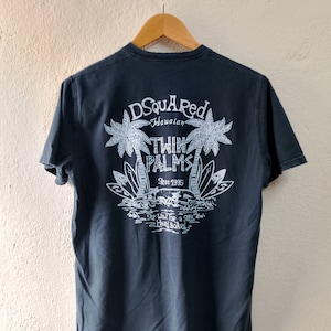 May include: A dark blue t-shirt with a white graphic print of palm trees and the text "D Squared Hawaiian Twin Palms Since 1985" and "Stay Wave for a Safe Hard Board".