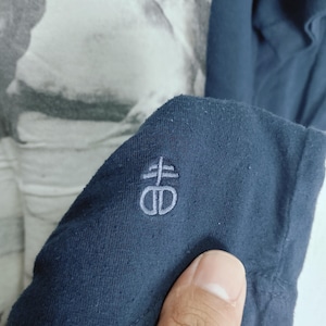 May include: Close-up of a navy blue long-sleeved shirt. The shirt features a light gray embroidered logo on the sleeve. The background shows a black and white graphic print.